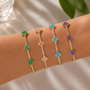 Puede incluir: Cuatro pulseras de cadena en tono dorado con pequeñas piedras transparentes brillantes y tres dijes en forma de trébol cada una. Los dijes son de color verde, rosa claro, azul brillante y morado, con cada pulsera mostrando un solo color.