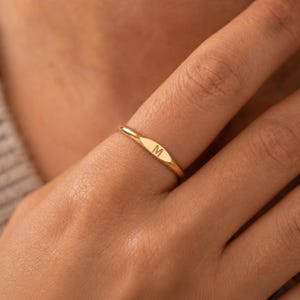Puede incluir: Un anillo fino de oro que presenta una pequeña placa plana de forma ovalada en la parte frontal. El centro de la placa está grabado con la letra mayúscula M. El aro del anillo es liso, pulido y mantiene un ancho fino y constante.