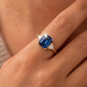 Puede incluir: Un anillo de oro con una gran piedra preciosa azul de corte esmeralda rectangular en el centro, flanqueada por dos piedras transparentes más pequeñas de corte triangular a cada lado. La banda es fina y pulida, con un diseño clásico.