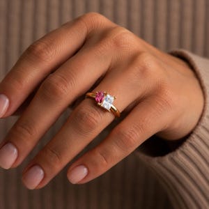 Puede incluir: Un anillo de oro con dos piedras preciosas distintas colocadas una al lado de la otra en una banda lisa. Una piedra es una gema rosa vibrante de corte ovalado, mientras que la otra es una piedra transparente de corte rectangular, fijadas con garras.