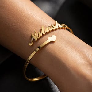 Puede incluir: Una pulsera de puño dorada con el nombre "Natasha" en escritura cursiva. La pulsera tiene un detalle en forma de corazón. La joya se muestra en una muñeca sobre un fondo oscuro.