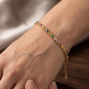 May include: A gold-toned bracelet featuring a series of small, round, multi-coloured gemstones set in individual bezels. The stones display a variety of hues including red, blue, green, yellow, and purple, arranged in a continuous line along the chain.