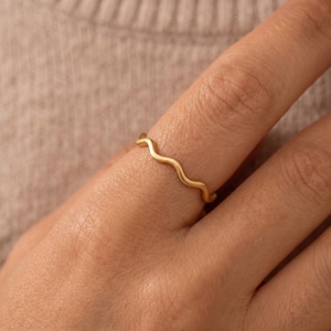 May include: A thin gold ring featuring a continuous wavy or serpentine design. The band has a smooth, polished finish and follows a gentle, undulating pattern that wraps around the finger, creating a simple and uniform aesthetic throughout.