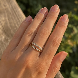 Puede incluir: Un anillo de oro con pequeñas piedras preciosas transparentes. El anillo se lleva en una mano con uñas cuidadas. El fondo es una vista borrosa de vegetación y una superficie de madera.
