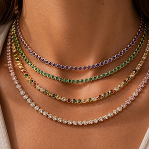 May include: Four layered gold-tone tennis necklaces featuring bezel-set round stones. The top necklace has purple stones, the second has green stones, the third has a mix of red, yellow, and green stones, and the bottom necklace has clear white stones.