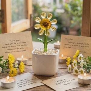 Puede incluir: Una escultura de flor de cristal con centro amarillo y pétalos transparentes sobre un tallo verde, plantada en una maceta cuadrada blanca con piedras pequeñas. El arreglo está rodeado por cuatro velas de té encendidas y notas escritas.