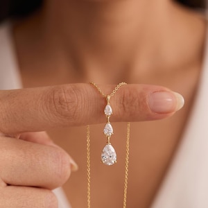 Puede incluir: Un collar de cadena dorada con un colgante vertical de tres piedras transparentes en forma de pera, dispuestas en orden descendente de tamaño. El colgante cuelga de una delicada cadena de eslabones dorados con acabado metálico pulido.