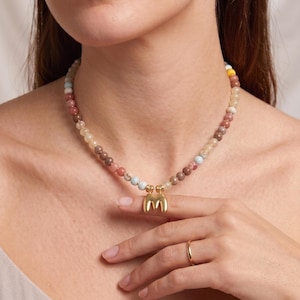 Puede incluir: Un collar de cuentas con una variedad de pequeñas piedras redondas y multicolores en tonos rosa, marrón, beige y azul claro, centrado con un colgante de metal dorado pulido en forma de letra mayúscula M.