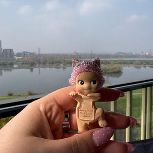 May include: A small, tan-colored figurine of a baby wearing a pink, glittery cat-ear hat. The figurine is held in a hand with pink nail polish. The background shows a body of water and a cityscape under a cloudy sky. The figurine has the text "Sandy Angel @DREAMS CHINA" on its back.