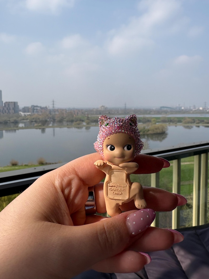 May include: A small, tan-colored figurine of a baby wearing a pink, glittery cat-ear hat. The figurine is held in a hand with pink nail polish. The background shows a body of water and a cityscape under a cloudy sky. The figurine has the text "Sandy Angel @DREAMS CHINA" on its back.