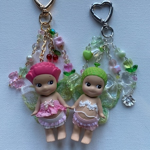 K&ouml;nnte beinhalten: Zwei kleine Feenfiguren mit rosa und gr&uuml;nen Akzenten, befestigt an Schl&uuml;sselanh&auml;ngern mit herzf&ouml;rmigen Verschl&uuml;ssen. Die Feen tragen rosa und wei&szlig;e Outfits und haben durchscheinende Fl&uuml;gel. Dekorative Ketten und Perlen in verschiedenen Farben h&auml;ngen an den Schl&uuml;sselanh&auml;ngern.