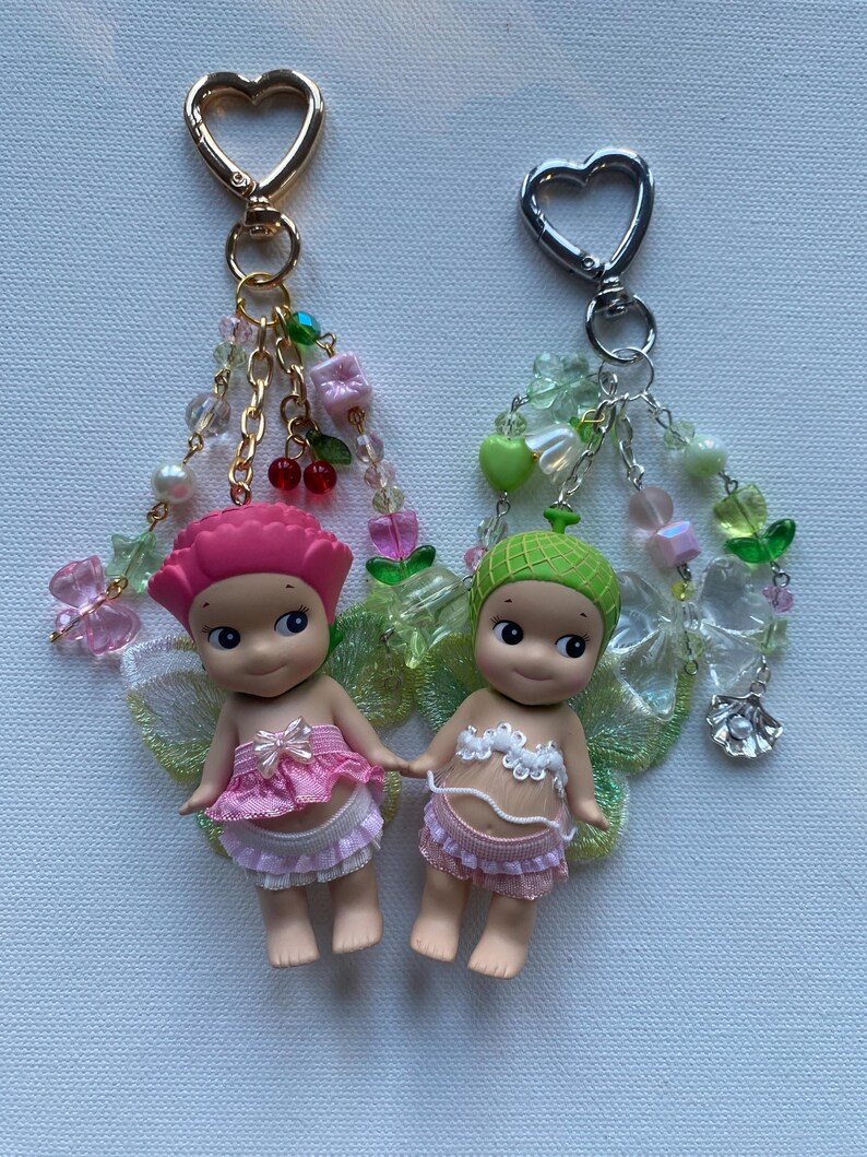 K&ouml;nnte beinhalten: Zwei kleine Feenfiguren mit rosa und gr&uuml;nen Akzenten, befestigt an Schl&uuml;sselanh&auml;ngern mit herzf&ouml;rmigen Verschl&uuml;ssen. Die Feen tragen rosa und wei&szlig;e Outfits und haben durchscheinende Fl&uuml;gel. Dekorative Ketten und Perlen in verschiedenen Farben h&auml;ngen an den Schl&uuml;sselanh&auml;ngern.