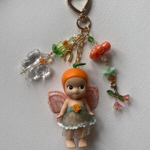 K&ouml;nnte beinhalten: Ein skurriles Schl&uuml;sselanh&auml;nger mit einer kleinen Puppe mit orangefarbenem Kopf und einem feenhaften Outfit. Die Puppe hat zarte Fl&uuml;gel und ist mit einem herzf&ouml;rmigen goldenen Verschluss, Ketten und dekorativen Anh&auml;ngern, darunter Kirschen und Blumen, verziert.