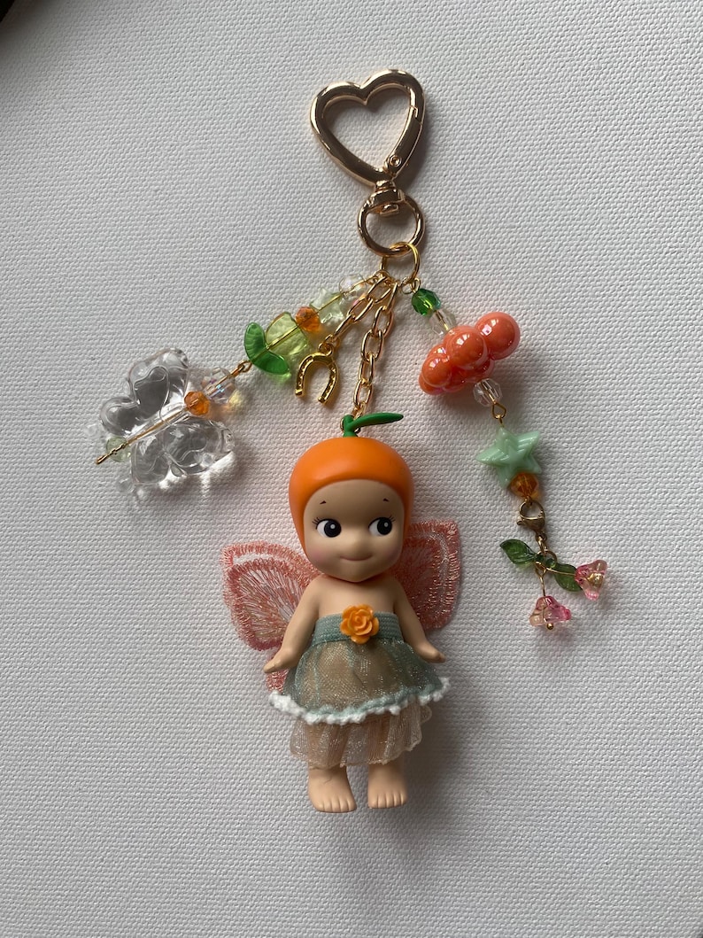 K&ouml;nnte beinhalten: Ein skurriles Schl&uuml;sselanh&auml;nger mit einer kleinen Puppe mit orangefarbenem Kopf und einem feenhaften Outfit. Die Puppe hat zarte Fl&uuml;gel und ist mit einem herzf&ouml;rmigen goldenen Verschluss, Ketten und dekorativen Anh&auml;ngern, darunter Kirschen und Blumen, verziert.