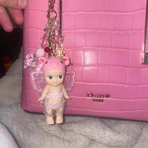 K&ouml;nnte beinhalten: Rosa Handtasche mit strukturierter Oberfl&auml;che, mit einem dekorativen Anh&auml;nger mit einer Ferkelfigur mit Krone und Fl&uuml;geln. Der Anh&auml;nger ist mit einer goldenen Kette befestigt und mit Perlen verziert.