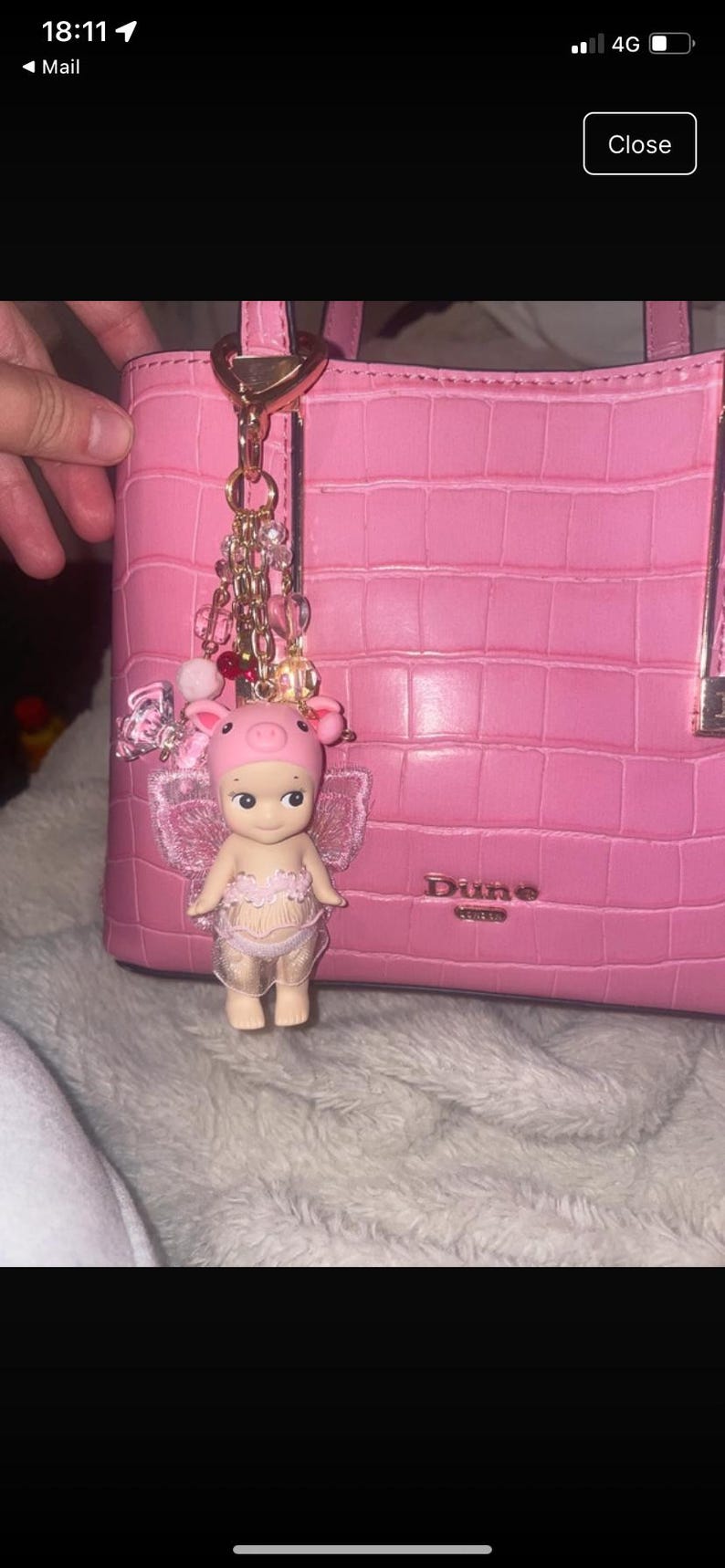 K&ouml;nnte beinhalten: Rosa Handtasche mit strukturierter Oberfl&auml;che, mit einem dekorativen Anh&auml;nger mit einer Ferkelfigur mit Krone und Fl&uuml;geln. Der Anh&auml;nger ist mit einer goldenen Kette befestigt und mit Perlen verziert.