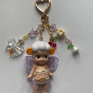 K&ouml;nnte beinhalten: Ein skurriler Schl&uuml;sselanh&auml;nger mit einer kleinen Puppe mit schaf&auml;hnlicher Frisur, orangefarbenen Ohren und lavendelfarbenen Fl&uuml;geln. Die Puppe tr&auml;gt ein durchsichtiges, spitzenbesetztes Outfit. Der Schl&uuml;sselanh&auml;nger hat einen herzf&ouml;rmigen goldenen Verschluss und dekorative Perlenstr&auml;nge mit Blumen und einem Schmetterling.