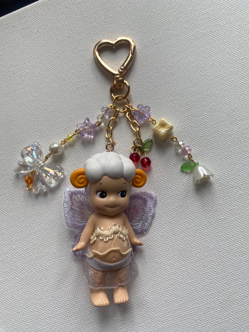 K&ouml;nnte beinhalten: Ein skurriler Schl&uuml;sselanh&auml;nger mit einer kleinen Puppe mit schaf&auml;hnlicher Frisur, orangefarbenen Ohren und lavendelfarbenen Fl&uuml;geln. Die Puppe tr&auml;gt ein durchsichtiges, spitzenbesetztes Outfit. Der Schl&uuml;sselanh&auml;nger hat einen herzf&ouml;rmigen goldenen Verschluss und dekorative Perlenstr&auml;nge mit Blumen und einem Schmetterling.