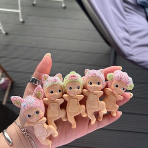 May include: A collection of small, baby-like figurines with pastel-colored hats and glitter accents. Each figurine has a unique hat design, including bunny ears and floral patterns. The figurines are held in a hand.