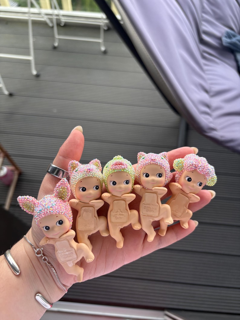 May include: A collection of small, baby-like figurines with pastel-colored hats and glitter accents. Each figurine has a unique hat design, including bunny ears and floral patterns. The figurines are held in a hand.