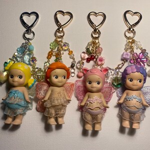 K&ouml;nnte beinhalten: Vier Schl&uuml;sselanh&auml;nger mit kleinen Puppenfiguren mit Fl&uuml;geln, jede mit einer anderen Haarfarbe und einem anderen Outfit. Die Schl&uuml;sselanh&auml;nger haben herzf&ouml;rmige Verschl&uuml;sse und Perlenakzente in verschiedenen Farben. Die Puppen haben einen s&uuml;&szlig;en, unschuldigen Ausdruck.