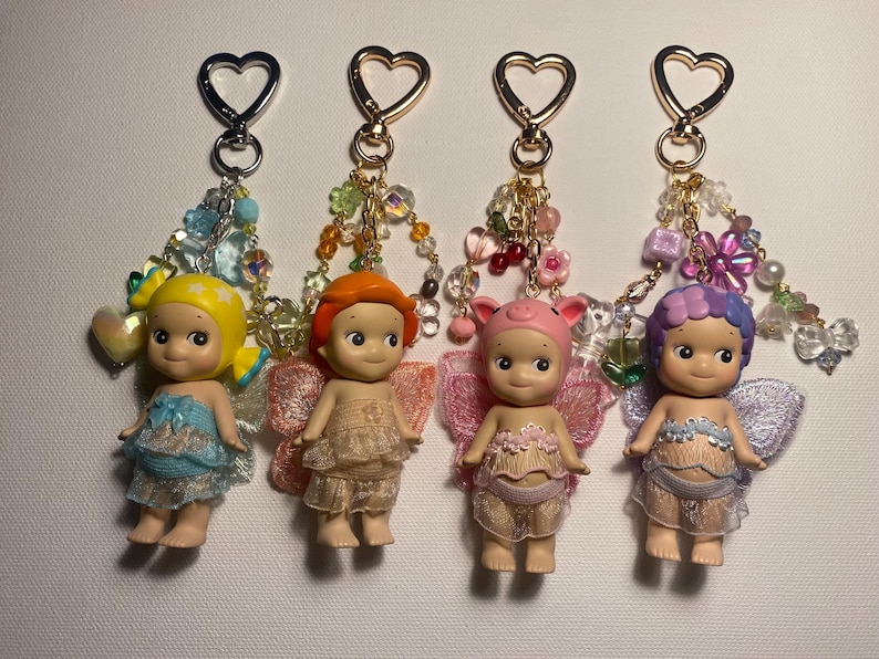 K&ouml;nnte beinhalten: Vier Schl&uuml;sselanh&auml;nger mit kleinen Puppenfiguren mit Fl&uuml;geln, jede mit einer anderen Haarfarbe und einem anderen Outfit. Die Schl&uuml;sselanh&auml;nger haben herzf&ouml;rmige Verschl&uuml;sse und Perlenakzente in verschiedenen Farben. Die Puppen haben einen s&uuml;&szlig;en, unschuldigen Ausdruck.