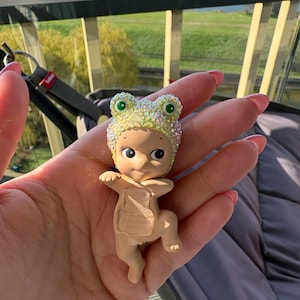 May include: A small figurine of a baby wearing a sparkly frog hat with green eyes. The baby is tan-skinned and has a neutral expression. The figurine is held in a hand, with a blurred background of a green landscape and a window.