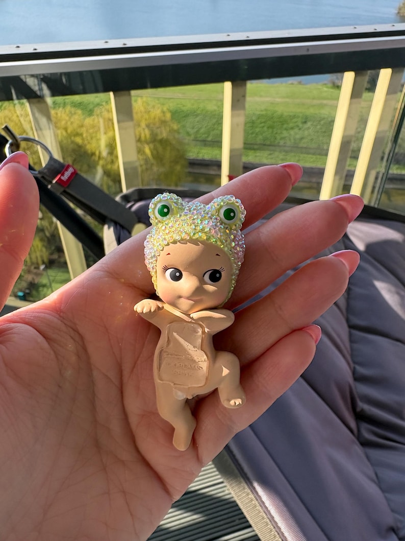 May include: A small figurine of a baby wearing a sparkly frog hat with green eyes. The baby is tan-skinned and has a neutral expression. The figurine is held in a hand, with a blurred background of a green landscape and a window.