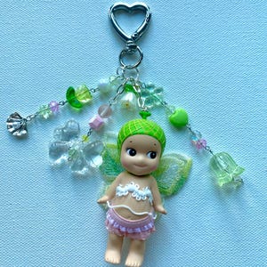 K&ouml;nnte beinhalten: Ein Schl&uuml;sselanh&auml;nger mit einer kleinen Puppe mit gr&uuml;ner M&uuml;tze, Feenfl&uuml;geln und einem rosa Rock. Die Puppe ist an einem silbernen herzf&ouml;rmigen Verschluss mit gr&uuml;nen, rosa und transparenten Perlen befestigt. Der Hintergrund ist hellblau.