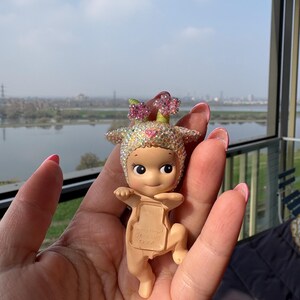 May include: A small, tan-colored figurine of a baby-like character with a flower crown and a jeweled head covering. The figurine is held in a hand, with a scenic background of a river and buildings visible.
