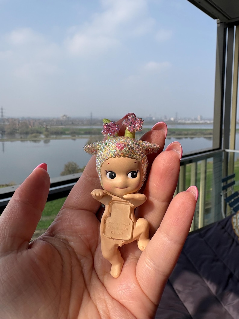 May include: A small, tan-colored figurine of a baby-like character with a flower crown and a jeweled head covering. The figurine is held in a hand, with a scenic background of a river and buildings visible.