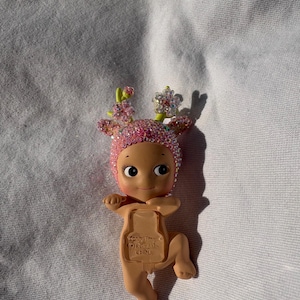 May include: A small, tan-colored baby doll with a pink, jeweled head covering and floral accents. The doll has a smiling face and is lying on a white surface. The doll's back has the text "(C)DREAMS CHINA".