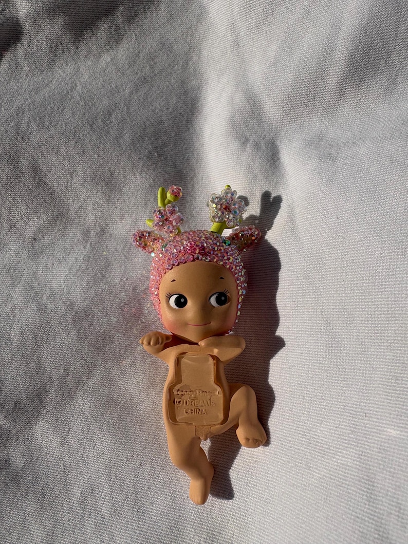 May include: A small, tan-colored baby doll with a pink, jeweled head covering and floral accents. The doll has a smiling face and is lying on a white surface. The doll's back has the text "(C)DREAMS CHINA".