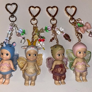K&ouml;nnte beinhalten: Vier Schl&uuml;sselanh&auml;nger mit kleinen Figuren mit Tierh&uuml;ten und Feenfl&uuml;geln. Jeder Schl&uuml;sselanh&auml;nger hat einen herzf&ouml;rmigen goldenen Verschluss und dekorative Charms. Die Figuren sind in verschiedenen Farben erh&auml;ltlich, darunter Blau, Beige und Rosa, mit detaillierten Outfits.