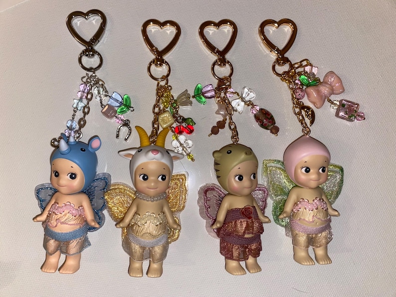 K&ouml;nnte beinhalten: Vier Schl&uuml;sselanh&auml;nger mit kleinen Figuren mit Tierh&uuml;ten und Feenfl&uuml;geln. Jeder Schl&uuml;sselanh&auml;nger hat einen herzf&ouml;rmigen goldenen Verschluss und dekorative Charms. Die Figuren sind in verschiedenen Farben erh&auml;ltlich, darunter Blau, Beige und Rosa, mit detaillierten Outfits.
