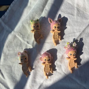 May include: Five small figurines with glittery hats in various colors, including pink, green, and purple, are arranged on a white surface. The figurines have a baby-like appearance with large eyes and outstretched arms. The hats feature animal-like designs.