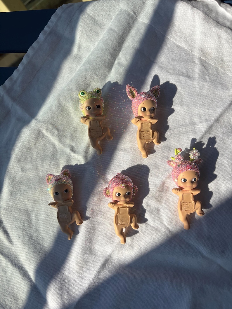 May include: Five small figurines with glittery hats in various colors, including pink, green, and purple, are arranged on a white surface. The figurines have a baby-like appearance with large eyes and outstretched arms. The hats feature animal-like designs.