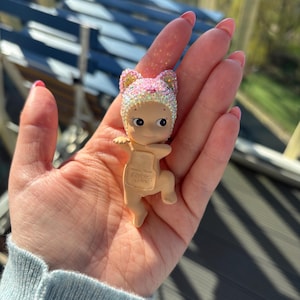 May include: A small, tan-colored figurine of a baby wearing a pink and gold glittery cat-eared hat. The figurine has large eyes and small arms and legs. The baby is holding a small, tan-colored backpack. The background is blurred.