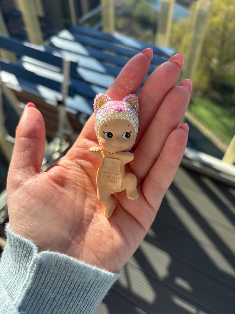 May include: A small, tan-colored figurine of a baby wearing a pink and gold glittery cat-eared hat. The figurine has large eyes and small arms and legs. The baby is holding a small, tan-colored backpack. The background is blurred.