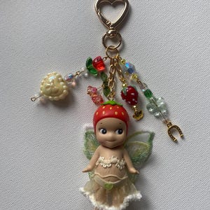 K&ouml;nnte beinhalten: Ein skurriler Schl&uuml;sselanh&auml;nger mit einer kleinen Feenpuppe mit Erdbeerm&uuml;tze und zarten Fl&uuml;geln. Die Puppe tr&auml;gt ein cremefarbenes Kleid und ein gr&uuml;nes Herz. Der Schl&uuml;sselanh&auml;nger hat einen herzf&ouml;rmigen goldenen Verschluss und Perlenanh&auml;nger.