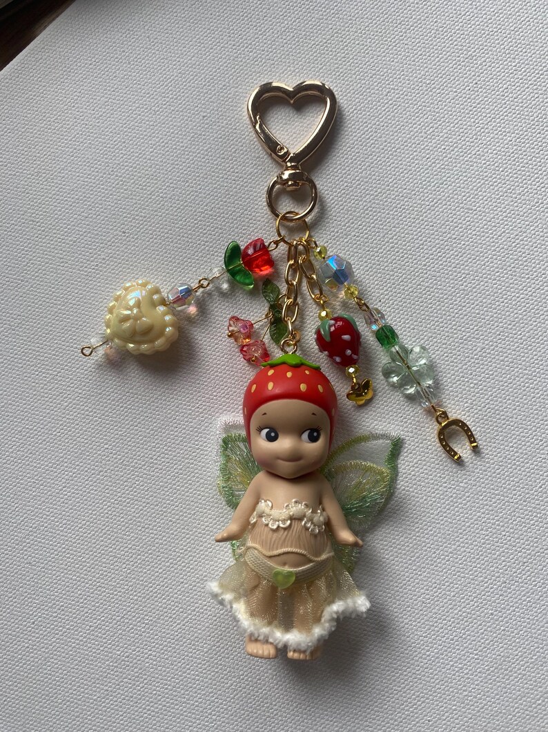 K&ouml;nnte beinhalten: Ein skurriler Schl&uuml;sselanh&auml;nger mit einer kleinen Feenpuppe mit Erdbeerm&uuml;tze und zarten Fl&uuml;geln. Die Puppe tr&auml;gt ein cremefarbenes Kleid und ein gr&uuml;nes Herz. Der Schl&uuml;sselanh&auml;nger hat einen herzf&ouml;rmigen goldenen Verschluss und Perlenanh&auml;nger.