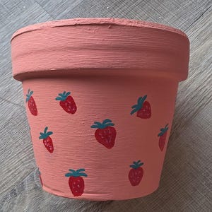 Puede incluir: Una maceta de color terracota con un diseño de fresas pintadas a mano. Las fresas son rojas con hojas verdes y están esparcidas por la maceta rosa. La maceta mide aproximadamente 15 cm de alto.