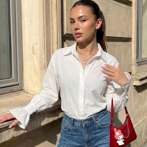 Puede incluir: Una camisa blanca de botones con mangas largas y pu&ntilde;os acampanados, combinada con pantalones vaqueros azules. Un peque&ntilde;o bolso de hombro rojo con cadena plateada y un dije de personaje de dibujos animados blanco y rosa.