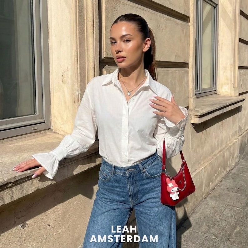 Puede incluir: Una camisa blanca de botones con mangas largas y pu&ntilde;os acampanados, combinada con pantalones vaqueros azules. Un peque&ntilde;o bolso de hombro rojo con cadena plateada y un dije de personaje de dibujos animados blanco y rosa.