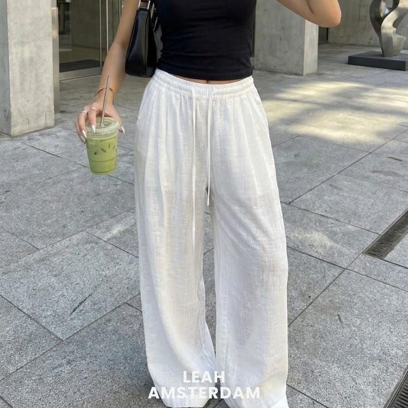 Peut inclure: Pantalon blanc &agrave; jambes larges et taille haute dot&eacute; d'une ceinture &eacute;lastique avec un cordon de serrage assorti. Le pantalon est confectionn&eacute; dans un tissu textur&eacute; et pr&eacute;sente une coupe ample et d&eacute;contract&eacute;e. Le texte LEAH AMSTERDAM est visible.