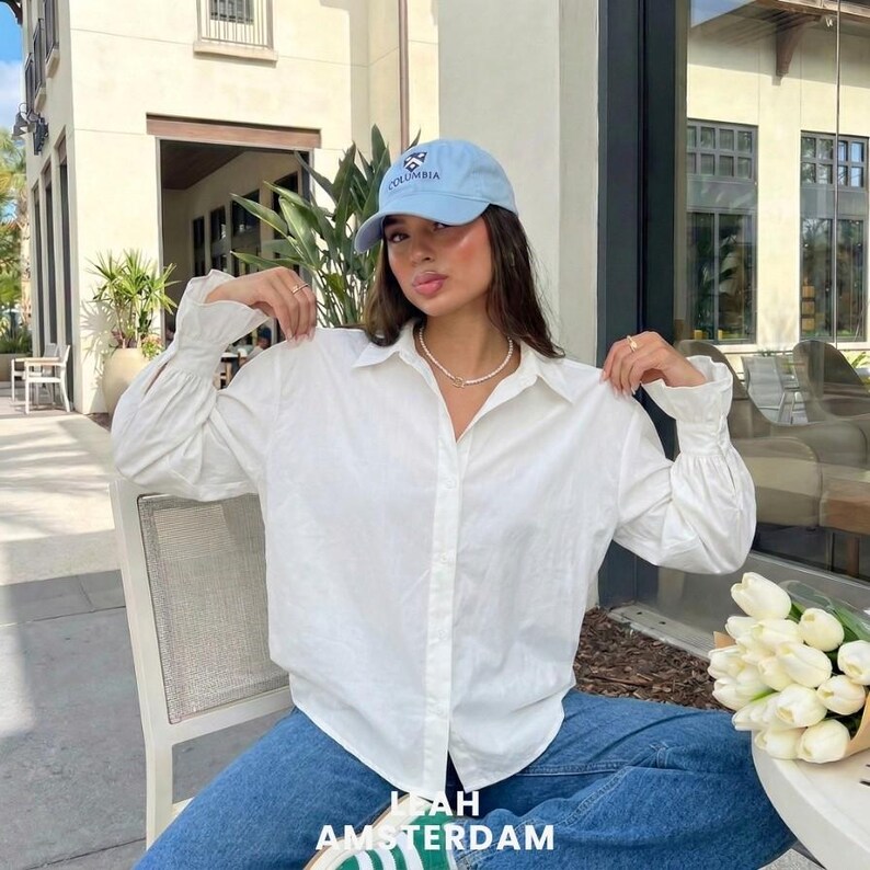 Puede incluir: Una gorra de b&eacute;isbol azul claro con la palabra Columbia y un logotipo de escudo bordados en la parte delantera, combinada con una camisa blanca de manga larga con botones, pu&ntilde;os fruncidos y cuello, usada con vaqueros azules y un collar de cuentas.