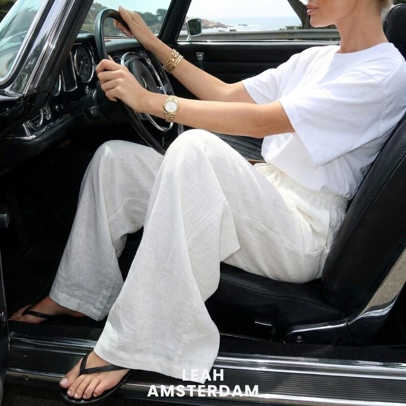 Peut inclure: Un t-shirt blanc &agrave; manches courtes et un pantalon blanc &agrave; jambes larges. La tenue est associ&eacute;e &agrave; des tongs noires. Le texte LEAH AMSTERDAM est visible dans la partie inf&eacute;rieure de l'image. Les v&ecirc;tements sont port&eacute;s dans une voiture vintage.