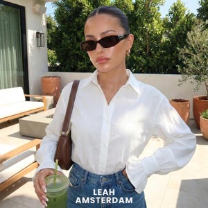 Puede incluir: Una camisa blanca de manga larga con botones, cuello y pu&ntilde;os acampanados, combinada con vaqueros azules, un bolso de hombro marr&oacute;n y gafas de sol rectangulares oscuras. El texto LEAH AMSTERDAM est&aacute; impreso en blanco en la parte inferior.