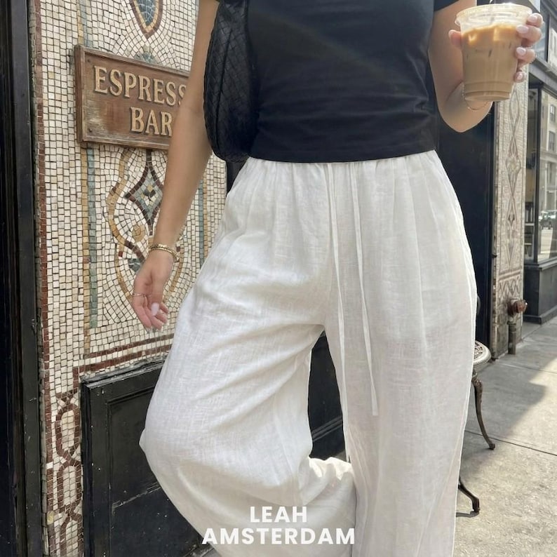 Peut inclure: Un t-shirt noir &agrave; manches courtes associ&eacute; &agrave; un pantalon blanc ample en lin dot&eacute; d'une taille &agrave; cordon de serrage. La tenue comprend un sac &agrave; bandouli&egrave;re noir tress&eacute; et un gobelet en plastique transparent contenant un caf&eacute; glac&eacute;.
