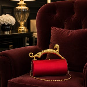 Puede incluir: Un bolso de mano cilíndrico de satén rojo con un asa de metal dorado en forma de leopardo estilizado. El bolso incluye una fina correa de cadena dorada y un pequeño cierre dorado en el borde superior del cuerpo cilíndrico.